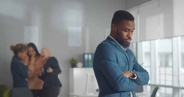 Portrait of Sad African Businessman Standing in Office with Female Colleagues Laughing at Him on alt