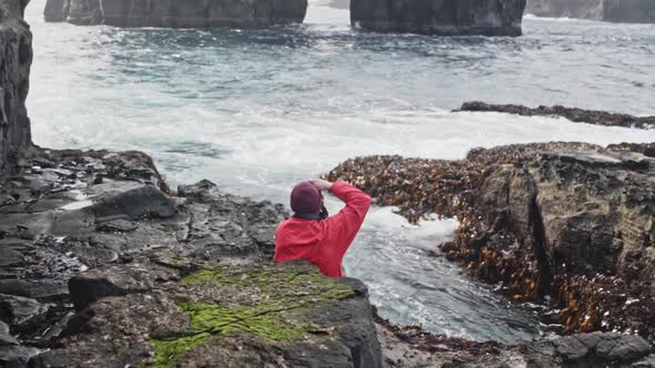 Man in Red Jacket Sitting on Rocks Taking a Picture of Sea Waves in Drangarnir alt