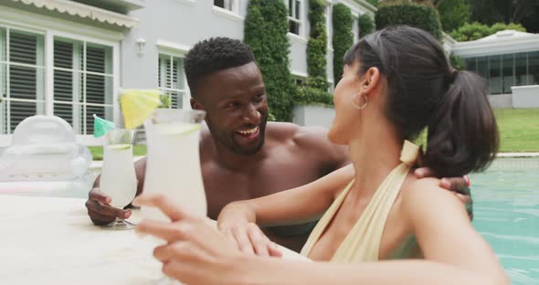 Happy diverse couple wearing swimming suits and drinking drinks at swimming pool in garden alt