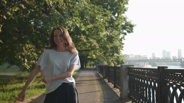a Woman in Casual Clothes is Dancing Merrily in a City Park Near an Openwork Lattice alt
