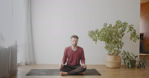 Caucasian Man Sitting Cross-legged Meditating with Closed Eyes Indoors in Sunny Apartment. Portrait