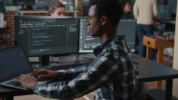 Portrait of African American Programer Typing on Laptop Sitting at Desk with Multiple Screens alt