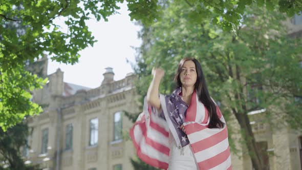 Beautiful Brunette Woman Wrapped in American Flag Raising Hand Up. Portrait of Confident Charming alt