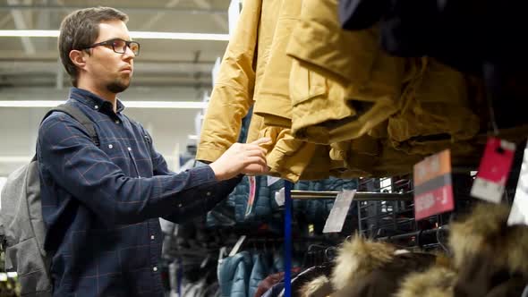 Adult Man is Looking and Touching Winter Jackets in a Sport Shop with Clothes alt