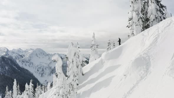 Snowboarder On Top Of Snowy Mountain Peak Looking Down The Ski Slope Aerial Drone Flying Past alt
