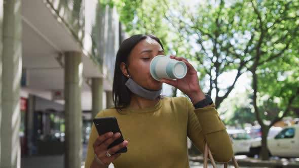 African american woman wearing face mask using smartphone drinking coffee in street alt
