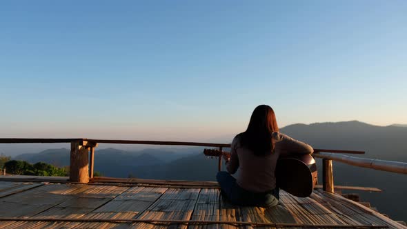 Rear view of a female sitting and playing guitar on wooden balcony