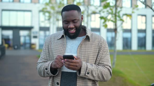 Happy AfricanAmerican Man in Casual Wear Using Smartphone Outdoors