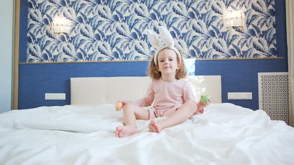 A Cute Little Girl with Long Hair Sits in Center of a Large Bed and Plays with Decorative Rabbit alt