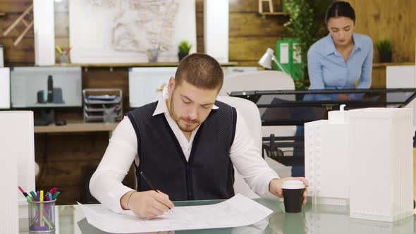 Tired Young Architect Taking a Sip of Coffee and Drawing a House Project alt