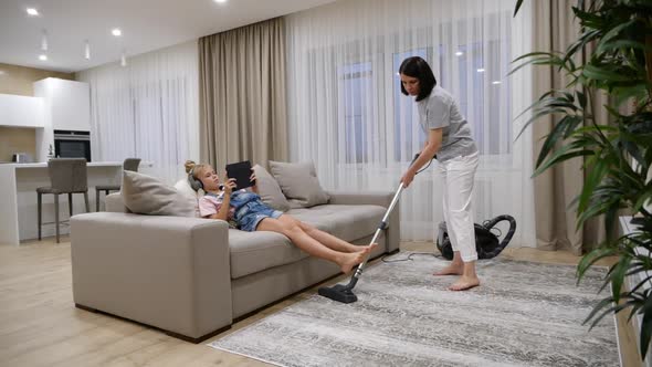 Mother Cleaning Floor in Living Room with a Vacuum Cleaner While Daughter Listen Music and Using
