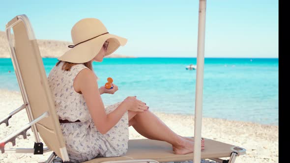 Woman in Hat Applies Sun Cream to Legs Relaxing on Deck Chair on Beach at Sea
