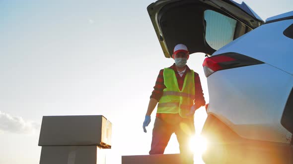 Delivery Man in Field in Overalls Loads Sealed Boxes with Product Purchases in Trunk Car alt