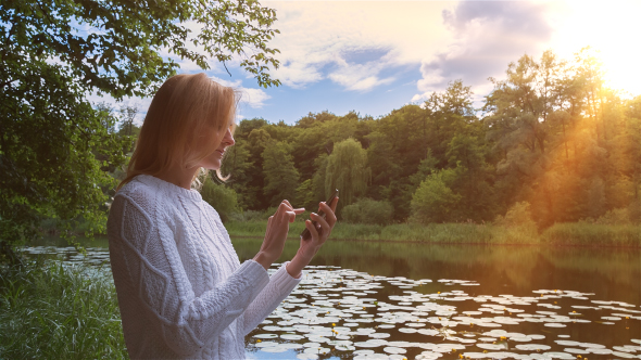 Beautiful Girl Using SmartPhone Sitting Near Lake in City Park 1 alt
