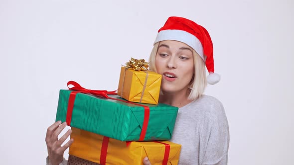 Young Pretty Blonde Girl in Christmas Hat Hardly Holding Stack of Falling Gift Boxes Over White alt