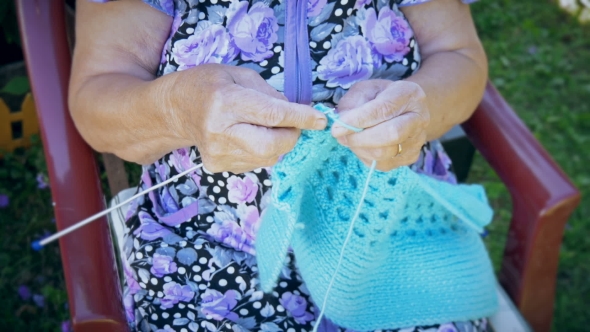 Elderly Woman Knitting Sitting On The Street Outside His Home.