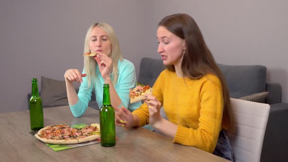 Two girls, drinking beer and eating delicious pizza, at home in their kitchen. alt