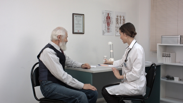 Smiling Pharmacist Holding a Bottle Of Pills And Pointing Them To Senior Patient. alt