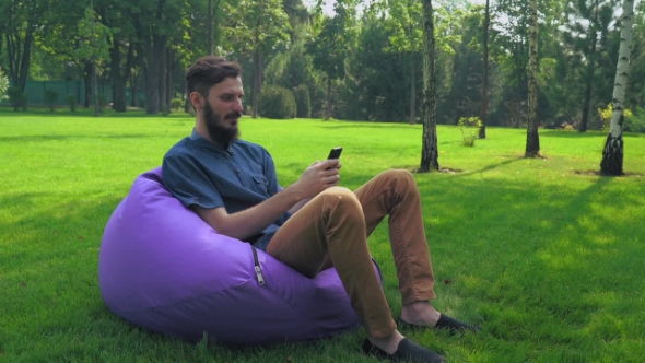 A Young Man In a Blue Shirt Sits On a Chair In The Park And Gaining a Message On The Phone. alt