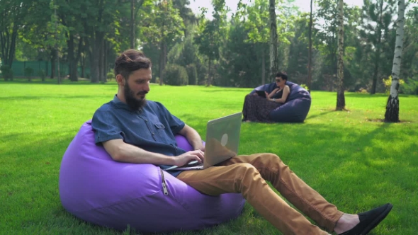 A Young Man In Blue Shirt Sitting In a Chair Ottoman Park And Working On The Computer. alt