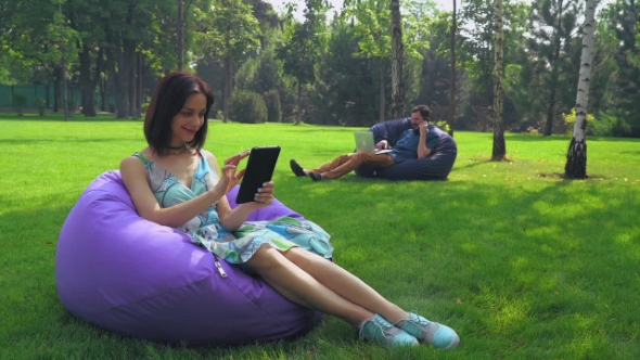 Young Brunette Girl Is Sitting In a Chair In The Park And Swipe Pages On Your Tablet. alt