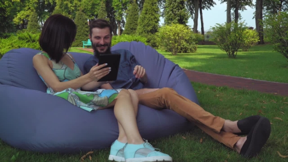 The Guy With The Girl Sit On a Chair And Ottoman Browsing Tablet. alt