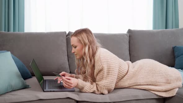Beautiful Blonde Girl Makes Online Payments with a Bank Card Using a Modern Laptop at Home Lying on alt