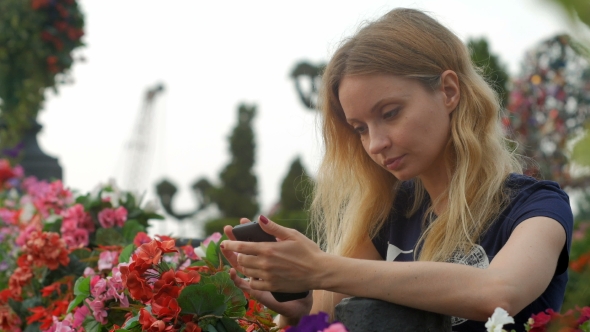 Girl With Long Wavy Blonde Hair Using Phone And Looking At Screen, Standing In Flower Bed