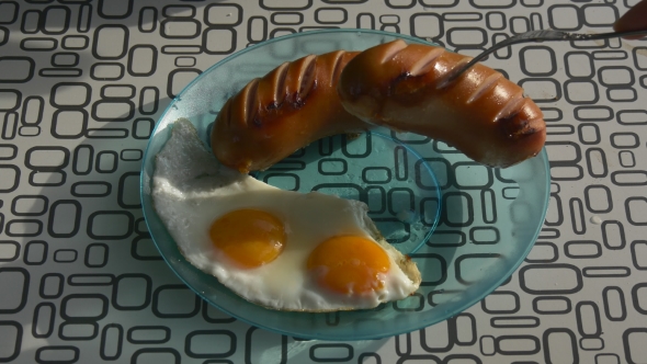Hand Puts Grilled Sausages On a Plate Near Fried Eggs