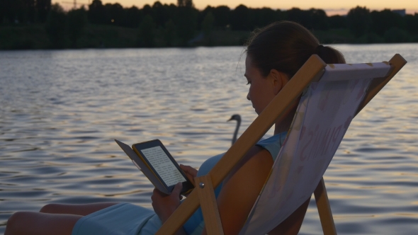 Girl Reads E-book At Sunset On The Sunbed Near Water