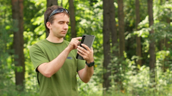 Young Man Touching Tablet In The Park Among The Trees. Green T-shirt And Sunglasses
