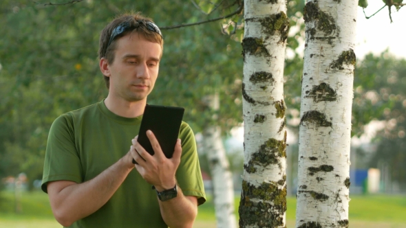 A Young Man Cyclist Touch Tablet Near The Birch . Against The Background Of Green Field 