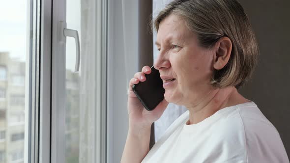Elderly Woman in White Tshirt Talks on Phone Near Window alt
