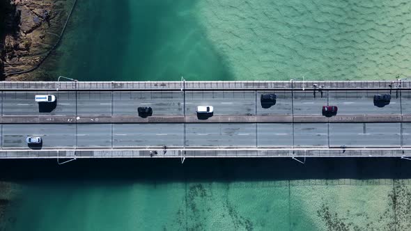 Motor vehicles and pedestrians travelling on a bridge spanning over a scenic ocean estuary. Panning alt