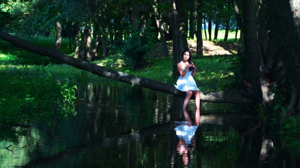 Young Brunette Girl Posing Near The Water, Sitting On a Fallen Tree.