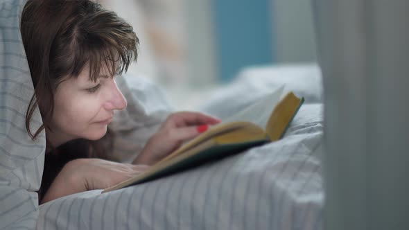 Woman Lies on the Bed and Reads a Book Covered with a Blanket