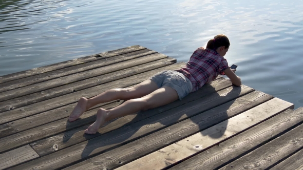 Woman Browses Mobile Phone Lying on a Wooden Pier