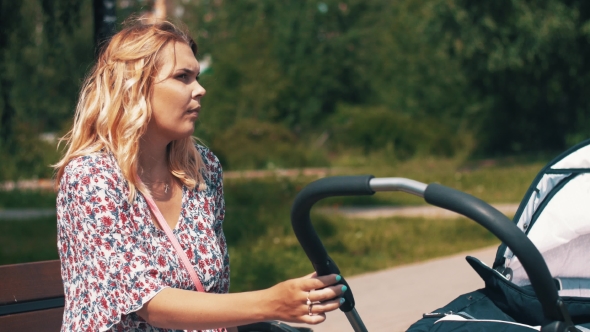 Young Mother Sway Baby Carriage Sitting On Bench At Park. Summer Sunny Day. alt