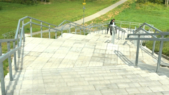 A Young Man Carries a Bicycle Through The Long Stairs. Against The Background Of Green Field