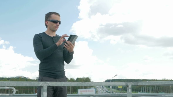 Outdoor Portrait Of Modern Young Man With Digital Tablet. A Man With Glasses On The Bridge