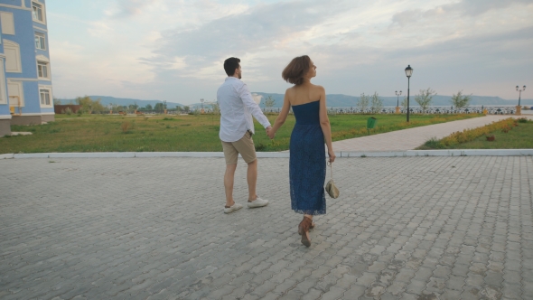 Beautiful Couple Enamored Young People Hold Hands And Walk Toward The Waterfront. alt