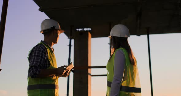 Builder Man with a Tablet and a Woman Inspector in White Helmets Shake Hands at Sunset Standing on alt