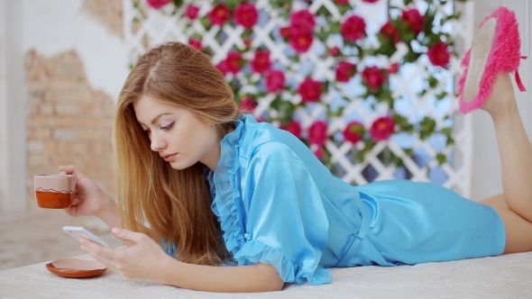 A Girl Is Lying On a Sofa, Drinking Coffee And Using a Smart Phone In The Morning