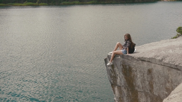 Young Attractive Backpacker Girl Relaxes By Resting On Mountain Rock Upon Lake Water Surface Below