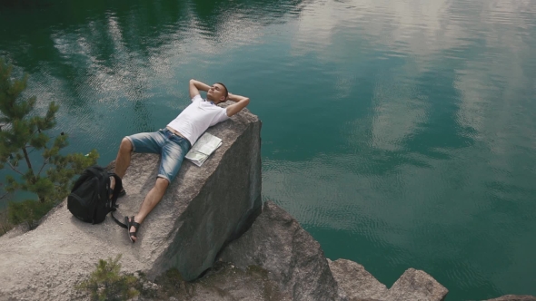 Young Tourist Man Relaxes By Resting On Rock Near Calm Mountain Lake Surface With Map Laying Beside