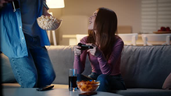 Young Woman Playing Video Game with Joystick Her Boyfriend Joining Her with Snacks and Drinks alt