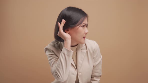 Asian business woman trying to hear something while standing isolated over beige background.