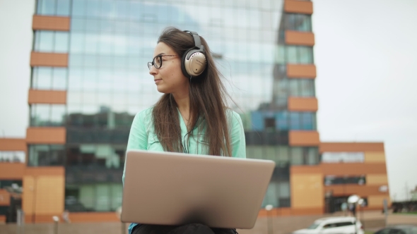 Attractive Young Girl With Headphones Sitting In The Open Space With a Laptop.