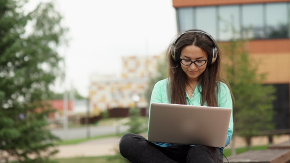 Attractive Young Girl With Headphones Sitting In The Open Space With a Laptop