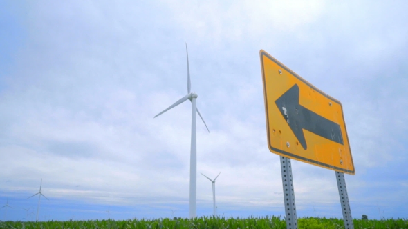 Road Sign Pointing To Wing Turbines Field Against Clouds Sky alt